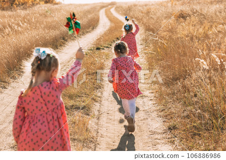 Playing in the wind: triplet girls run towards adventures with windmills on a warm autumn day, using wind energy 106886986