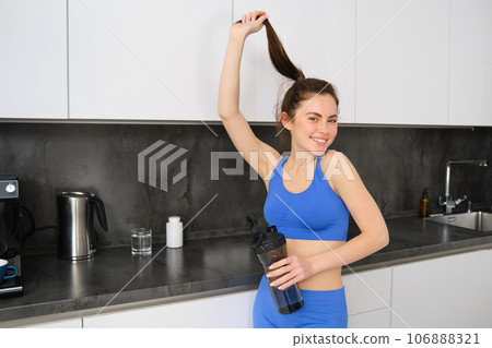 Portrait of smiling, beautiful young woman drinking water and dietary supplement, taking vitamins after workout, showing her strong, healthy and shiny hair Portrait of smiling, beautiful young woman drinking water and dietary supplement, taking vitamins after workout, showing her strong, healthy and shiny hair 106888321