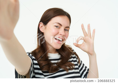 Portrait of positive, happy young woman shows okay sign and takes selfie, holds smartphone with extended hand, posing near something she recommends, isolated on white background 106888325