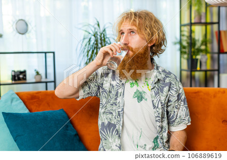 Thirsty Caucasian man sits at home holding glass of natural aqua make sips drinking filtered water Thirsty Caucasian man sits at home holding glass of natural aqua make sips drinking filtered water 106889619