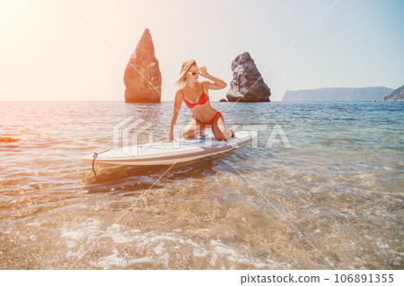 Woman sea sup. Close up portrait of happy young caucasian woman with blond hair looking at camera and smiling. Cute woman portrait in red bikini posing on sup board in the sea Woman sea sup. Close up portrait of happy young caucasian woman with blond hair looking at camera and smiling. Cute woman portrait in red bikini posing on sup board in the sea 106891355