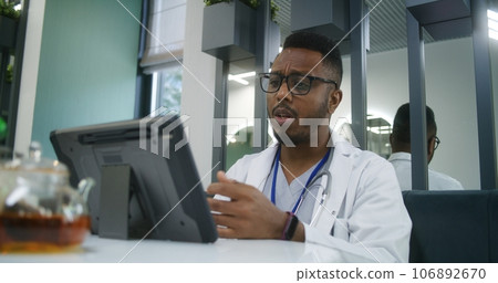Doctor sits in hospital cafeteria, has online appointment on digital tablet 106892670