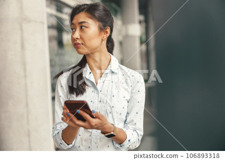 Female entrepreneur looking at side while holding smartphone standing on modern office background 106893318