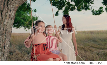 Positive mother rides cute little girls on swings in summer park at twilight time. Little girls play swings with loving mother in country field. Mother amuses little daughter on swings in garden Positive mother rides cute little girls on swings in summer park at twilight time. Little girls play swings with loving mother in country field. Mother amuses little daughter on swings in garden 106893549