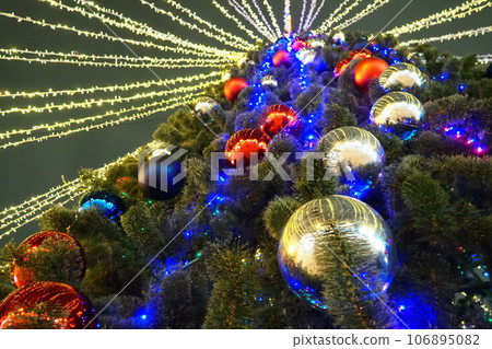 Multi-colored glass shiny balls on the New Year's city tree. Glowing garlands hang over the Christmas tree. Outdoor winter decoration. Festive design of the city square. Red, blue and yellow ball Multi-colored glass shiny balls on the New Year's city tree. Glowing garlands hang over the Christmas tree. Outdoor winter decoration. Festive design of the city square. Red, blue and yellow ball 106895082
