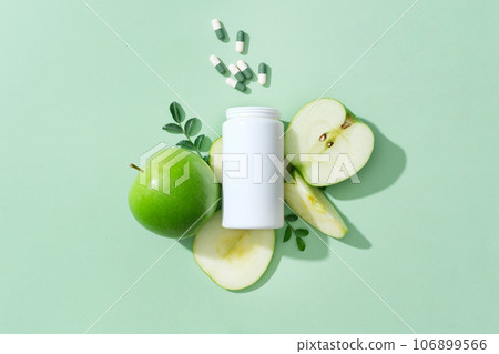 On the pastel background, a white medicine bottle unlabeled with capsule pills decorated with fresh slices of green apple. Advertising photo for product with natural extract for weight loss 106899566