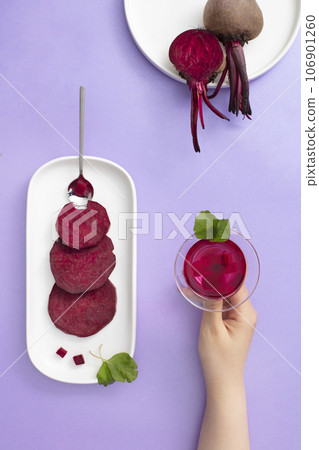 Top view of ceramic dishes containing slices of beetroot decorated with glass cup of beetroot juice on hand's woman on purple background. Beets are rich in antioxidants and essential minerals Top view of ceramic dishes containing slices of beetroot decorated with glass cup of beetroot juice on hand's woman on purple background. Beets are rich in antioxidants and essential minerals 106901260