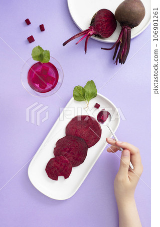 On the purple background, fresh slices of beetroot displayed on white dishes, decorated with glass cup of beetroot juice. Woman's hand holding a spoon holding a small piece of beetroot  106901261