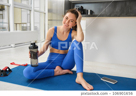 Portrait of beautiful sportswoman in blue sportswear, drinks water, stays hydrated during fitness training session, smiling at camera Portrait of beautiful sportswoman in blue sportswear, drinks water, stays hydrated during fitness training session, smiling at camera 106901656