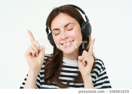 Portrait of happy student girl, listening music in headphones, dancing and pointing fingers at copy space, promo text, isolated against white background 106901659