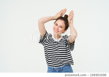 Cute young woman shows animal ears gesture above head, smiling and looking happy, posing in casual clothes over white background 106901696