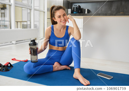 Smiling fitness girl drinks water in the middle of training session, smiling and looking happy, workout from home in blue sportsbra Smiling fitness girl drinks water in the middle of training session, smiling and looking happy, workout from home in blue sportsbra 106901739