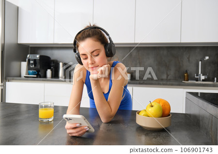 Image of happy, stylish young sports woman, standing in kitchen and drinking orange juice, listening music in headphones, using smartphone app 106901743
