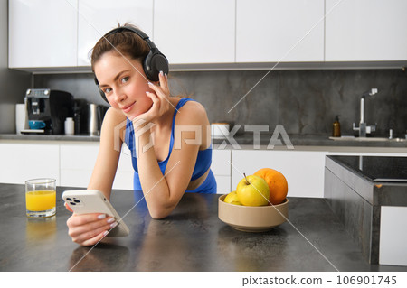Portrait of young fitness woman with headphones, drinking orange juice in kitchen and using smartphone, listening music, getting ready for workout gym 106901745