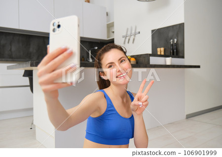 Close up portrait of beautiful brunette sportswoman, taking selfie on smartphone, posing on fitness mat with mobile phone, records her fitness exercises, workout training 106901999