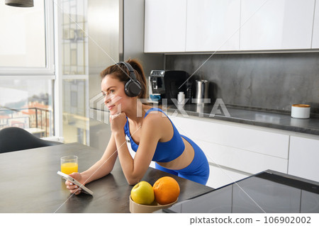 Portrait of stylish sportswoman, listening music in headphones, looking at smartphone, watching video on mobile phone, standing in kitchen, wearing activewear Portrait of stylish sportswoman, listening music in headphones, looking at smartphone, watching video on mobile phone, standing in kitchen, wearing activewear 106902002