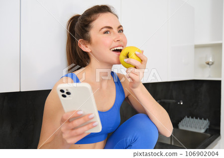 Portrait of stylish, young fitness woman, eating an apple and using mobile phone, holding smartphone, wearing sportswear 106902004