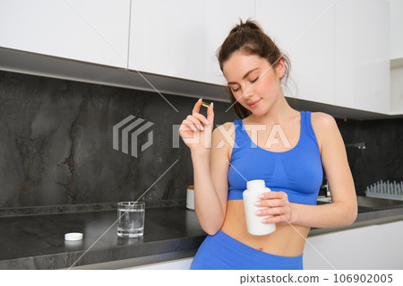 Image of young brunette woman with slim, fit body, holding dietary supplements, vitamin B, fish oil, standing in kitchen 106902005