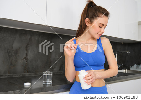 Image of young brunette woman with slim, fit body, holding dietary supplements, vitamin B, fish oil, standing in kitchen 106902188