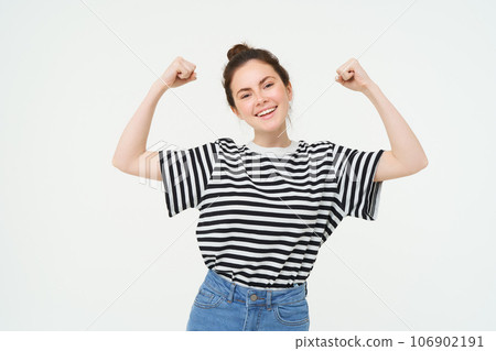 Woman power and feminism. Young girl feeling empowered and strong, flexing her biceps, showing muscles on arms, standing over white background 106902191