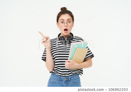 Portrait of girl gasps in surprised, points left with amazed face. Shocked student shows something, pointing finger at banner, holding notebooks, white background Portrait of girl gasps in surprised, points left with amazed face. Shocked student shows something, pointing finger at banner, holding notebooks, white background 106902203