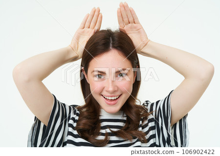 Image of cute brunette girl shows animal eats with hands on top of head, smiling and looking happy, white background 106902422