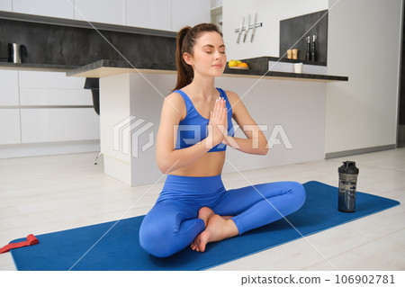 Sport and recreation. Close up portrait of beautiful girl does yoga at home, clasps her hands and meditates, sits on rubber mat in kitchen, makes asana, relaxing Sport and recreation. Close up portrait of beautiful girl does yoga at home, clasps her hands and meditates, sits on rubber mat in kitchen, makes asana, relaxing 106902781