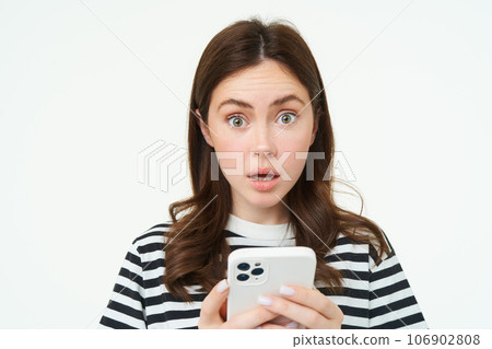 Portrait of woman with smartphone in hands, looking surprised, raising eyebrows from amazement, white background 106902808