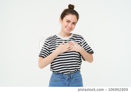 Young smiling woman, feels gratitude, thankful for something, holds hands on heart and looks tender at camera, standing over white background 106902809
