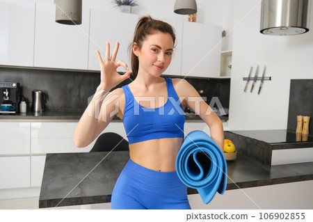 Portrait of stylish fitness instructor, young fit woman, shows okay sign, holds yoga mat in hand, approves workout smartphone app, aerobics training at home 106902855