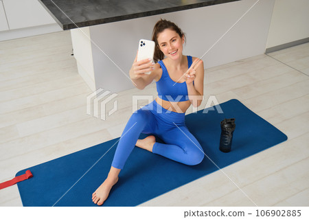 Portrait of stylish, smiling young brunette woman, wears blue sportswear, poses on yoga mat with smartphone, shows peace sign and takes selfie, does workout at home Portrait of stylish, smiling young brunette woman, wears blue sportswear, poses on yoga mat with smartphone, shows peace sign and takes selfie, does workout at home 106902885