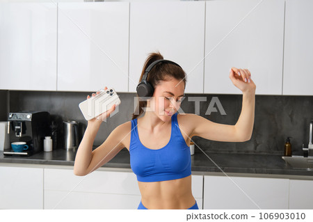 Portrait of beautiful fitness blogger, woman in headphones, listening music and dancing in kitchen, wearing blue leggings and sportsbra 106903010