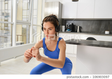 Image of young sportswoman, fitness instuctor doing squats workout at home, listening music in wireless headphones and stretching arms, doing exercises in living room 106903097