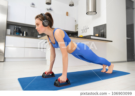 Portrait of woman in wireless headphones, doing workout from home, standing in plank with sports equipment, listening music, looking confident 106903099