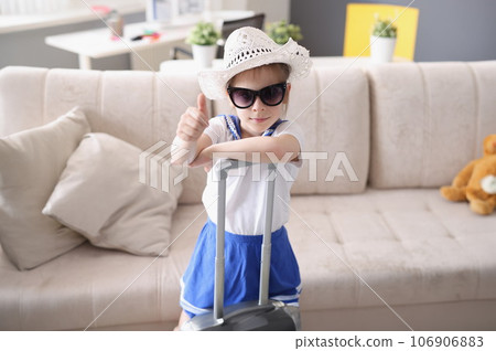 Little girl in hat and sunglasses stands with suitcase and holds thumbs up 106906883