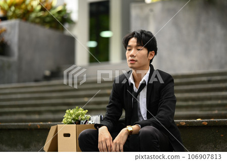 Disappointed male worker sitting on the stairs of building with box of personal stuff. Dismissal from work, unemployment concept 106907813