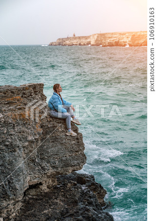 woman sea travel. A woman in a blue jacket sits on a rock above a cliff above the sea, looking at the stormy ocean. Girl traveler rests, thinks, dreams, enjoys nature woman sea travel. A woman in a blue jacket sits on a rock above a cliff above the sea, looking at the stormy ocean. Girl traveler rests, thinks, dreams, enjoys nature 106910163