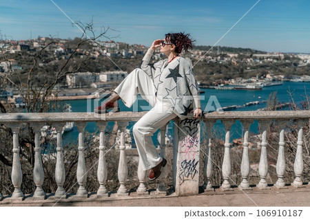 Woman walks around the city, lifestyle. A young beautiful woman in white trousers and a sweater sits on a white fence with balusters and overlooks the sea bay and the city. Woman walks around the city, lifestyle. A young beautiful woman in white trousers and a sweater sits on a white fence with balusters and overlooks the sea bay and the city. 106910187