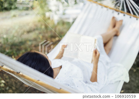 A female brunette is lying in a hammock outdoors and reading a book. Blurred background. 106910559