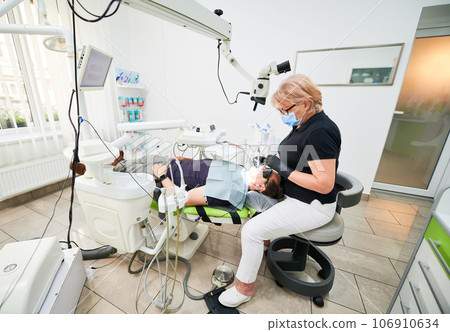 Side view of female dentists sitting on chair, fixing teeth. Doctor inspecting, examining problem, holding equipment, wearing uniform, gloves and mask. Concept of dentistry. Side view of female dentists sitting on chair, fixing teeth. Doctor inspecting, examining problem, holding equipment, wearing uniform, gloves and mask. Concept of dentistry. 106910634