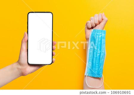 One woman's hand is holding a medical mask, while the other hand is holding a cellphone. Mock up. Yellow background. Concept of protection against coronavirus and other diseases One woman's hand is holding a medical mask, while the other hand is holding a cellphone. Mock up. Yellow background. Concept of protection against coronavirus and other diseases 106910691