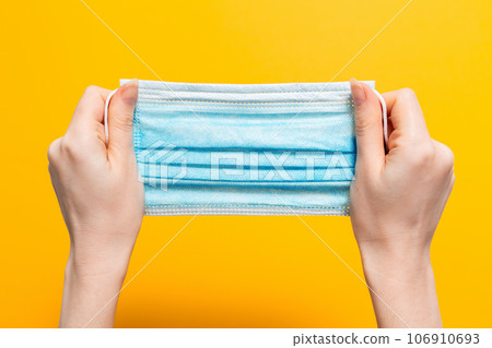 Female's hands hold tight medical mask. Yellow background. Close up. Concept of protection against coronavirus and other diseases Female's hands hold tight medical mask. Yellow background. Close up. Concept of protection against coronavirus and other diseases 106910693