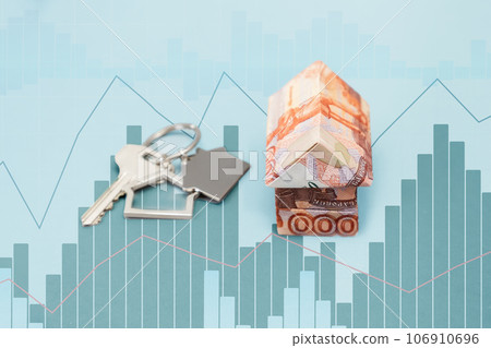 Close up of paper origami house made of Russian rubles. Light blue background with charts. The concept of mortgages and purchase of housing 106910696