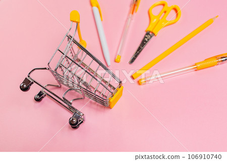 Concept of back to school and shopping. Toy shopping cart with yellow office supplies on pink background Concept of back to school and shopping. Toy shopping cart with yellow office supplies on pink background 106910740
