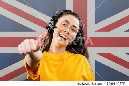 World English Language Day. Portrait of a young smiling woman wearing headphones, giving a thumbs-up. On the background of the English flag. The concept of learning foreign languages World English Language Day. Portrait of a young smiling woman wearing headphones, giving a thumbs-up. On the background of the English flag. The concept of learning foreign languages 106910761