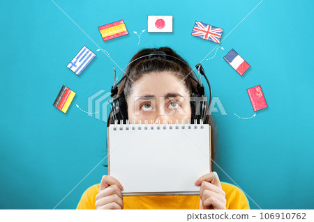 Portrait of a young woman wearing headphones, holding a notebook and looking up at the flags of different countries, covering half of her face. The concept of learning foreign languages Portrait of a young woman wearing headphones, holding a notebook and looking up at the flags of different countries, covering half of her face. The concept of learning foreign languages 106910762