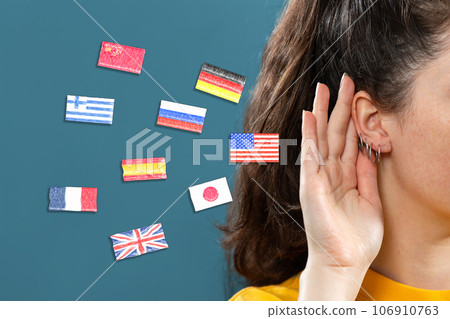 Woman put her hand to her ear, into which the flags of different countries fly. Close up. Blue background. The concept of learning foreign languages Woman put her hand to her ear, into which the flags of different countries fly. Close up. Blue background. The concept of learning foreign languages 106910763