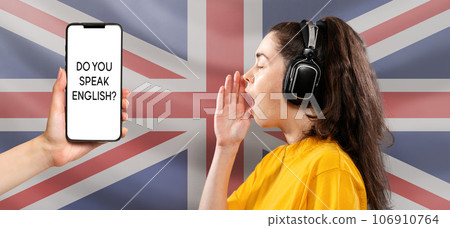 Online language learning. Portrait of a young woman wearing headphones, her hand over her open mouth. English flag on the background. Female hand holding a cellphone. Concept of English language day Online language learning. Portrait of a young woman wearing headphones, her hand over her open mouth. English flag on the background. Female hand holding a cellphone. Concept of English language day 106910764