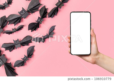 A woman's hand holds a smartphone. In the left corner is a flock of bats made of paper. Purple background. Mock up. The concept of Halloween and technologies A woman's hand holds a smartphone. In the left corner is a flock of bats made of paper. Purple background. Mock up. The concept of Halloween and technologies 106910779