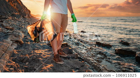 A male volunteer holding two polyethilene bag with garbage. In the background the sea and the sunset. Banner with copy space. The concept of environmental protection and coastal clean up A male volunteer holding two polyethilene bag with garbage. In the background the sea and the sunset. Banner with copy space. The concept of environmental protection and coastal clean up 106911025